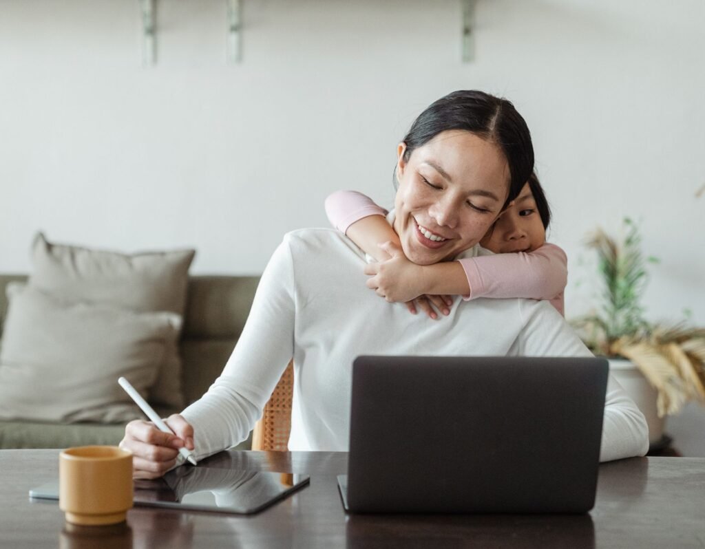 mom doing work on laptop, toddler behind her hugging her around the neck