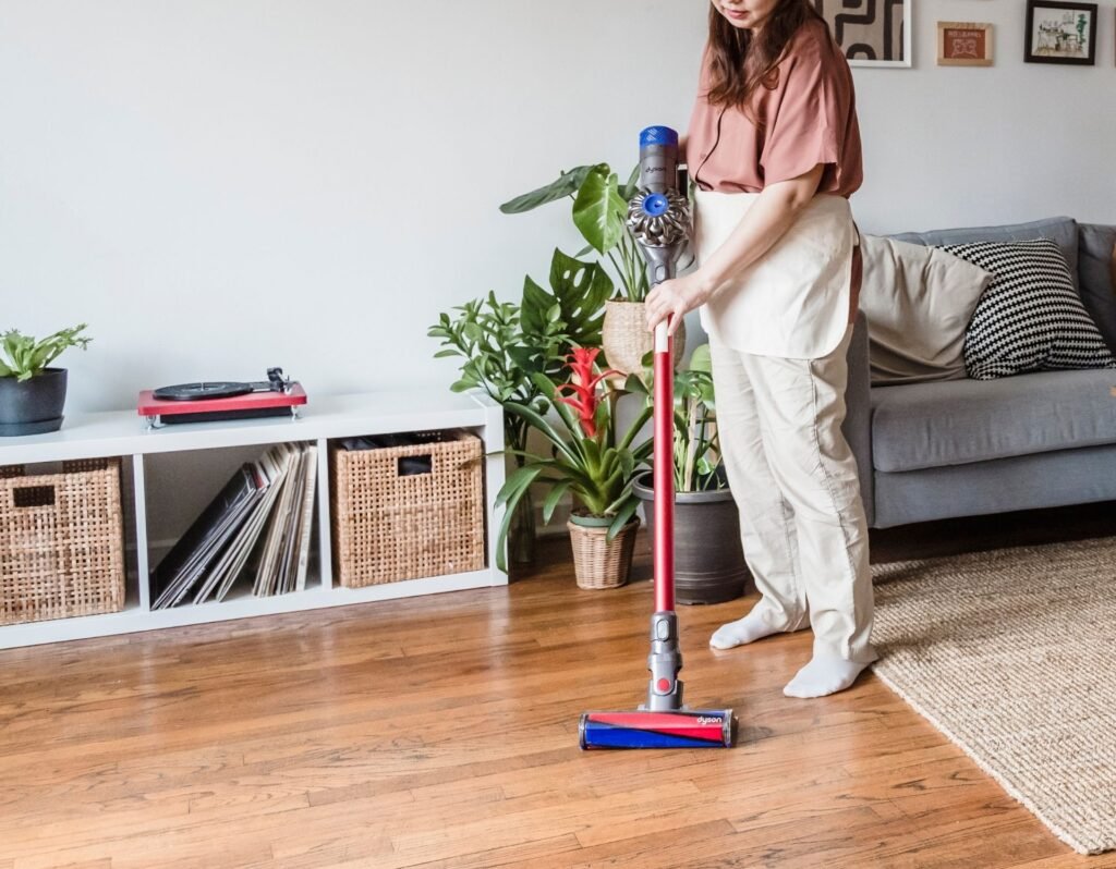 Woman holding a cordless vacuum cleaning tool, cleaning the hard wood floors with an organized shelf, plants and a couch behind her. 