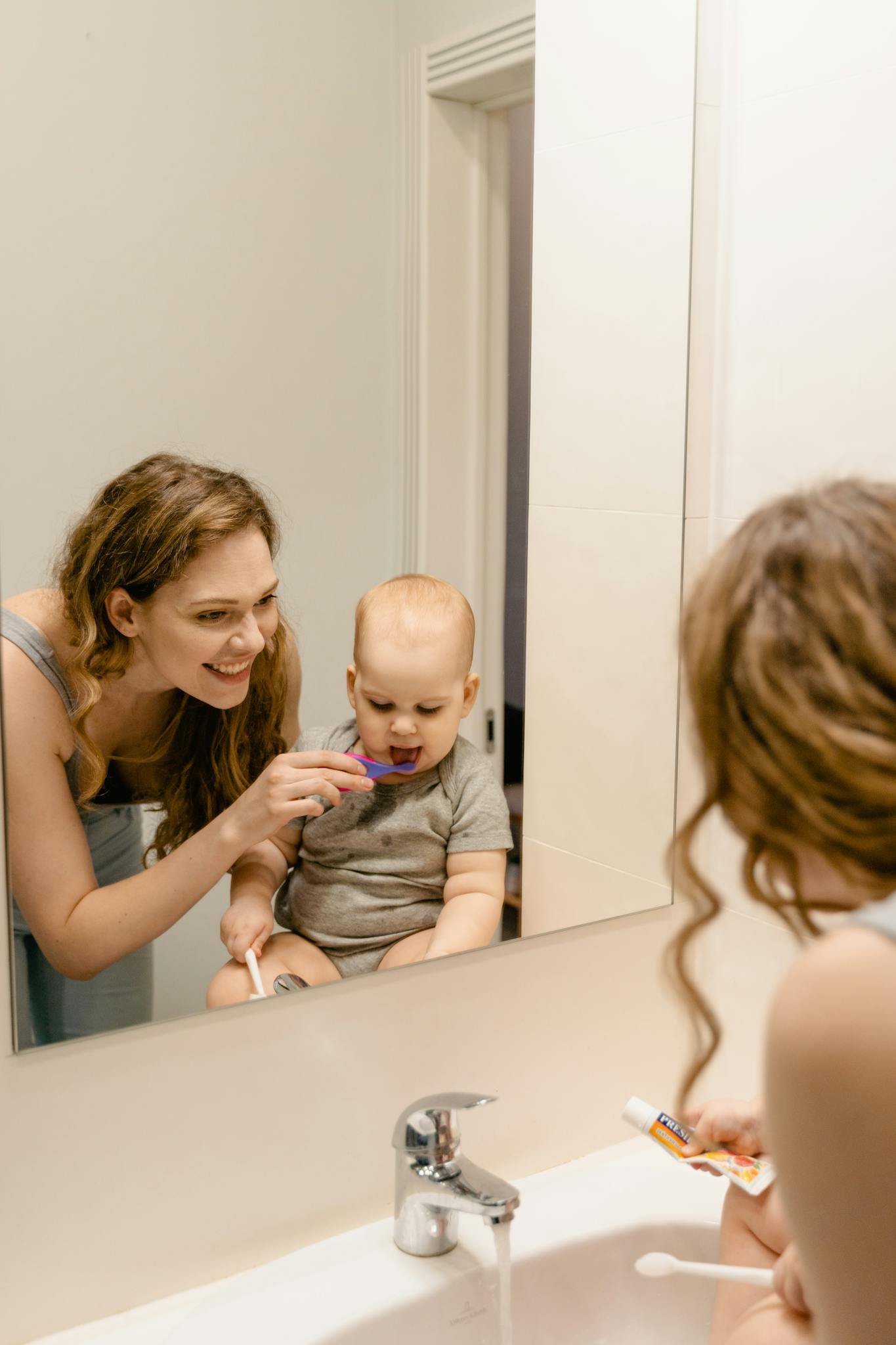 A mother helps her baby brush teeth in the home bathroom mirror, ensuring good hygiene habits.