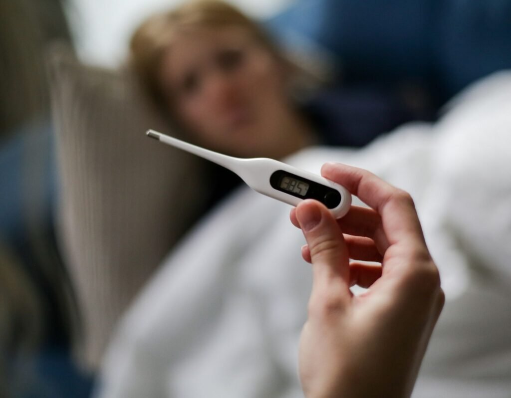 thermometer showing a fever temperature with woman blurred in the background laying in bed resting while sick